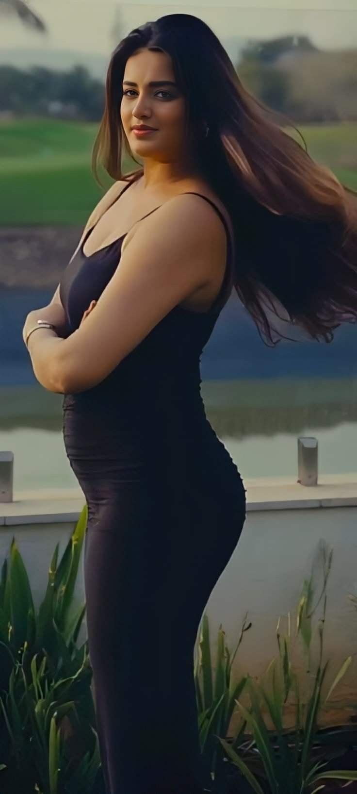 a woman with long hair standing in front of a body of water wearing a black dress