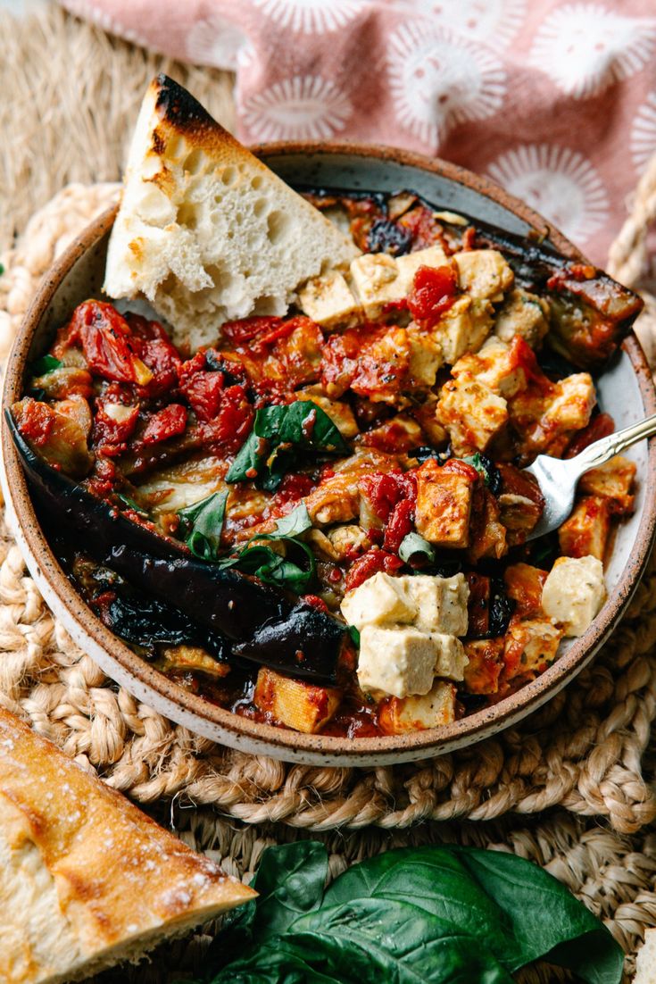 a bowl filled with food on top of a woven table cloth next to bread and greens