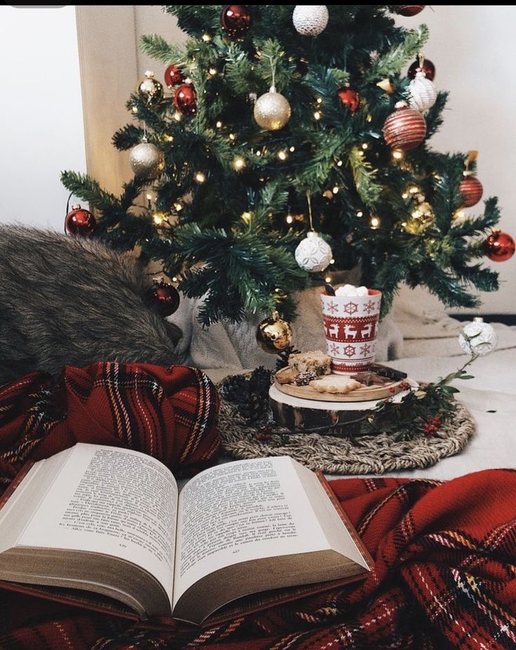 an open book sitting on top of a blanket next to a christmas tree with ornaments