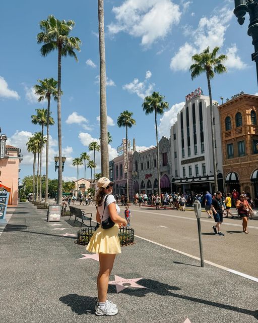 a woman is standing on the sidewalk in front of some palm trees and people are walking around