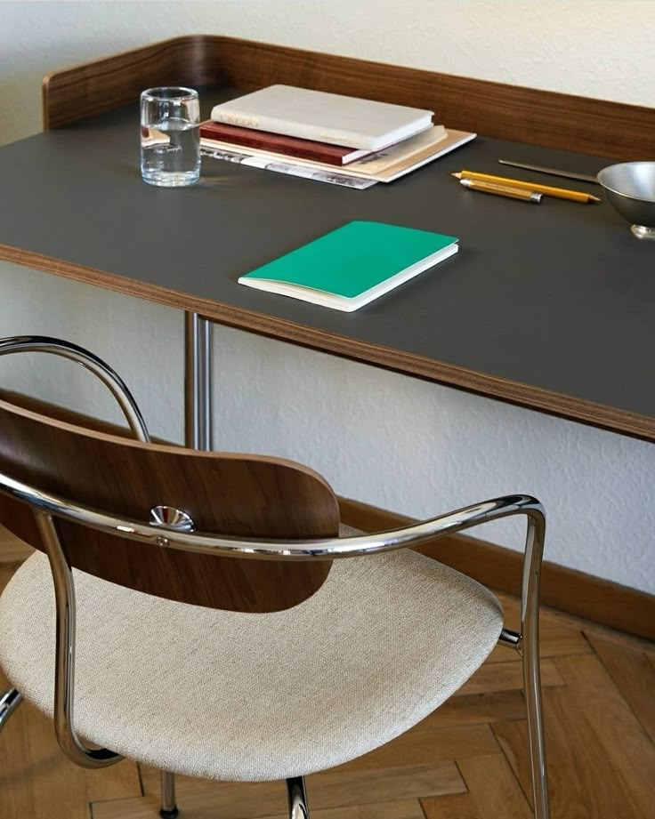 a desk with a chair, notebooks and water glass on it in front of a white wall