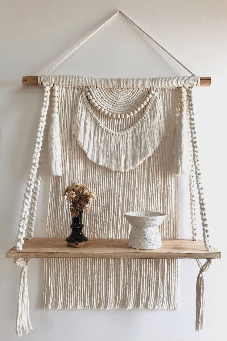 a white wall hanging with some tassels on top of it and a vase filled with flowers