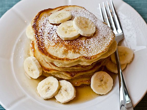 pancakes with bananas and powdered sugar on a white plate next to silverware, fork and knife