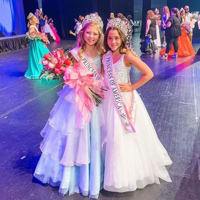 two girls in dresses and tiaras posing for the camera on stage with flowers around their necks