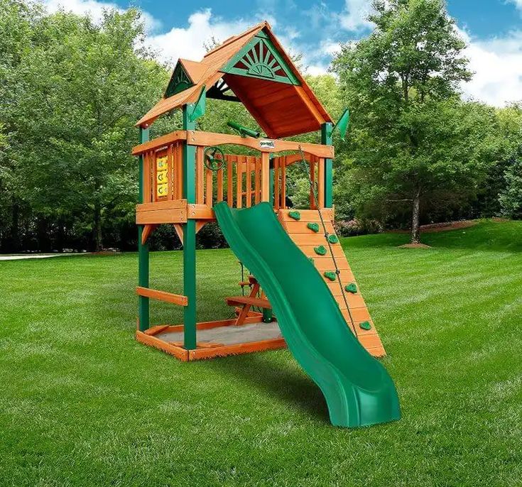 a wooden playset with a green slide and trees in the backgrouds