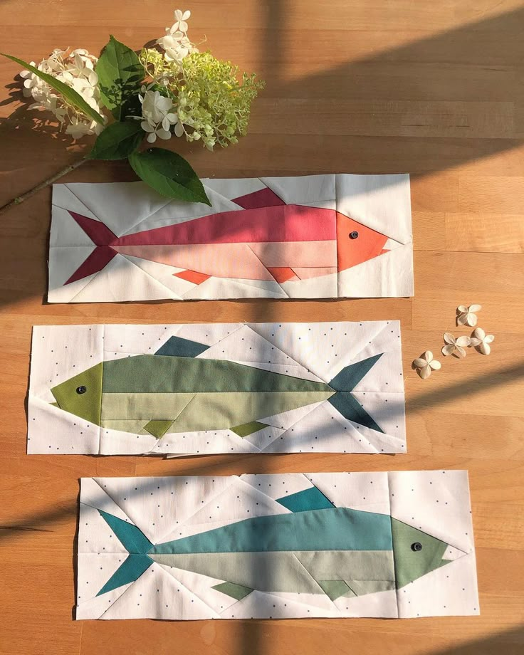 three pieces of paper with fish on them sitting on a wooden table next to flowers