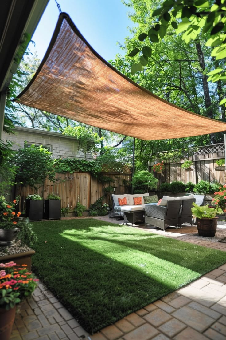 Cozy backyard patio with wicker furniture, pillows, and a shade sail overhead. Surrounded by greenery, flowers, and wooden fencing. Backyard Shading Ideas, Backyard Shade Sails Ideas, Garden Shade Cloth Ideas, Creating Shade In Backyard, Garden Shade Sail, Cozy Backyard Patio, Backyard Shade Ideas, Home Owners Association, Canvas Canopy