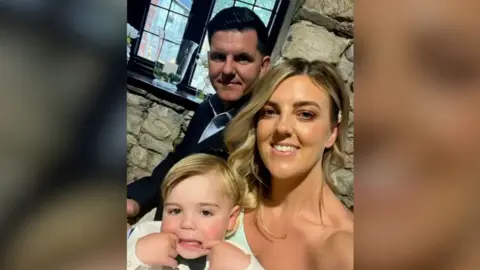 Ryan and Victoria Thomas, with their little boy in front of them who is blonde and has his fingers in his mouth, pulling a silly face