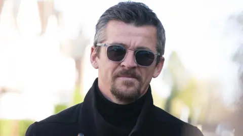 PA Media Joey Barton, who has greying hair swept to the right and a goatee beard, looks into the camera wearing a black woollen style coat over a black  turtle-neck jumper. 