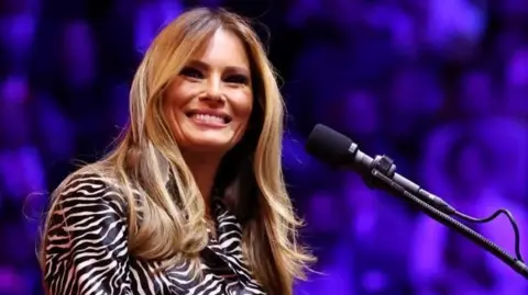 Melania Trump on stage at Madison Square Garden, with a microphone and a purple background