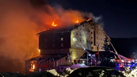 The exterior of the Grand Kartal Hotel at night with the roof in flames and two ladders from fire trucks extended.