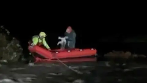 A man in a hi-vis in a red boat in a flooded field. There is also a man with his dog in the boat.