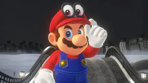 Nintendo Screenshot shows Mario in a world from Super Mario Odyssey, looking enigmatically at the viewer as he tweaks the brim of his famous red cap with a white-gloved hand.