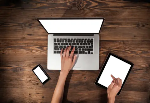 Getty Images A laptop, a phone and a tablet are on a wooden table. Two white hands are shown typing and working on the tablet. 