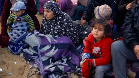 Palestinians wait to be allowed to return to their homes in northern Gaza after they were displaced to the south at Israel's order during the war. A woman in a headscarf sits with a blanket on her knees, with a child wearing a red jumper next to her.