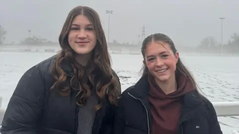 L-R Millie and Zara pictured at a snow-covered rugby pitch. The girls have long brown hair worn loose and wear black puffer coats. The sky behind them is grey. 