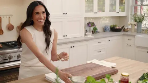 Netflix A promotional photo released from the With Love, Meghan programme shows the smiling duchess in a bright and spacious kitchen. She wears a sleeveless cream top and skirt.