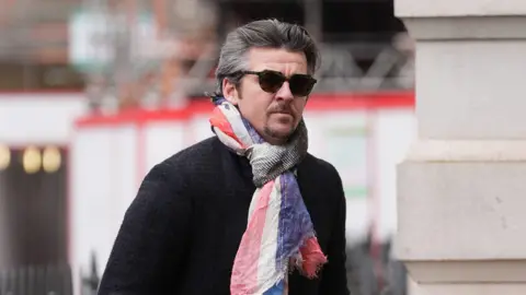 PA Media Joey Barton arriving at Westminster Magistrates' Court on 24 January. Mr Barton, who has grey hair and a goatee, is wearing sunglasses, a red white and blue scarf and a dark tweed coat