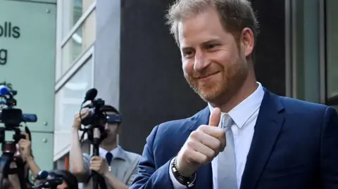 Reuters Prince Harry, dressed in a blue suit and grey tie, gives a thumbs up to supporters as he leaves the High Court's Rolls Building in 2023, with media cameras in the background, during his evidence against the Mirror Group titles who he was suing at the time for unlawful intrusion. 