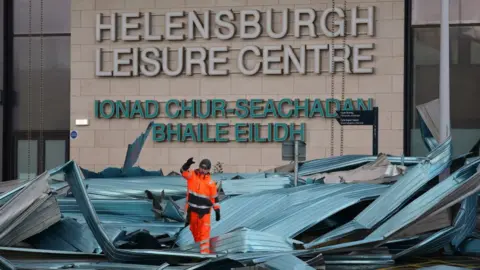 A man in orange overalls is walking on top of the metal roof of the leisure centre and the roof is in the car park, badly damaged.