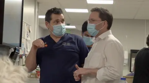 A BBC reporter, wearing a white shirt and a mask, is speaking to a nurse, also wearing a mask and blue hospital scrubs