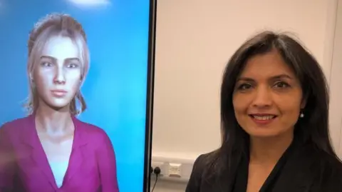 A woman is wearing a black suit, she has long dark hair she is standing next to a large screen with a female avatar on it wearing a purple shirt 