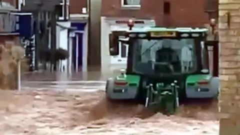 Tractor driving through flood water in Tenbury Wells in a flooded village street with metres-high water 