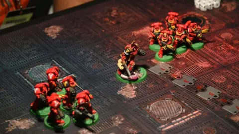 Getty Images The top of a gaming table with 11 Space Marine miniatures on it. The characters hold large gun-like weapons and their suits of futuristic armour are carefully painted in the same deep red colour with gold accents for small details. In the corner a clutch of several dice can be seen.