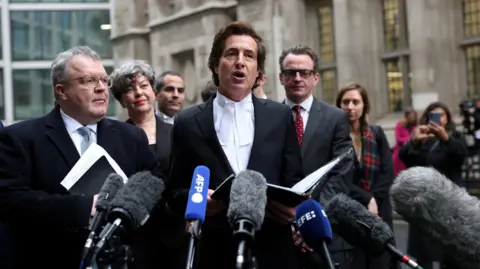 Reuters David Sherborne, wearing bands, speaking to reporters at the High Court flanked on the left by Lord Tom Watson, the former Labour MP. Several other people stand behind them and there are several reporters' microphones in the foreground.