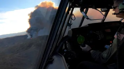 A large cloud of orange smoke is seen through the window of a plane with a pilot inside.