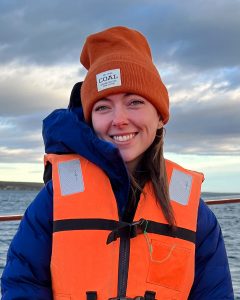 Portrait photo of McKenzie. She's wearing a life vest and wooly hat and is at sea.