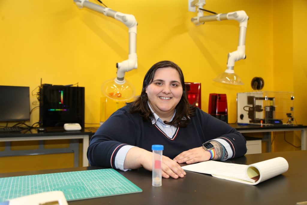 Photo of Sara sitting at a table in a lab. There are robotic arms attached to the wall behind her.