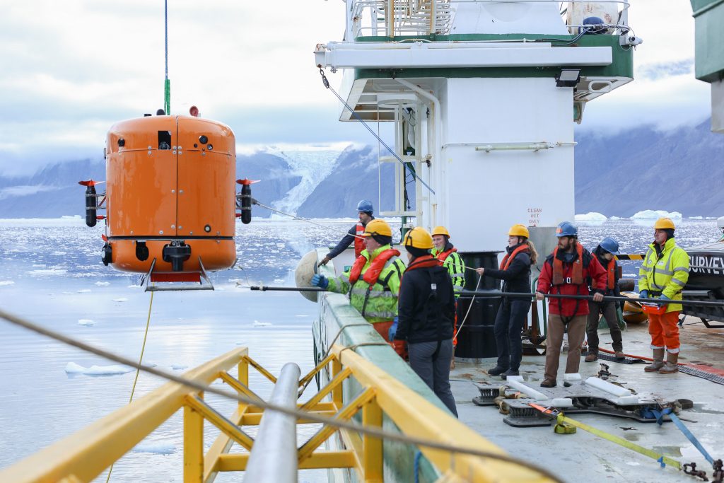 People in hard hats on the deck of a ship guiding the robot submersible back on deck.