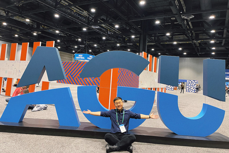 Photo of a person sitting in front of the AGU sign in the convention center with their arms outstretched.