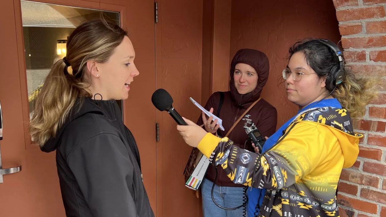 Young Indigenous woman interviews woman in front of brick building.