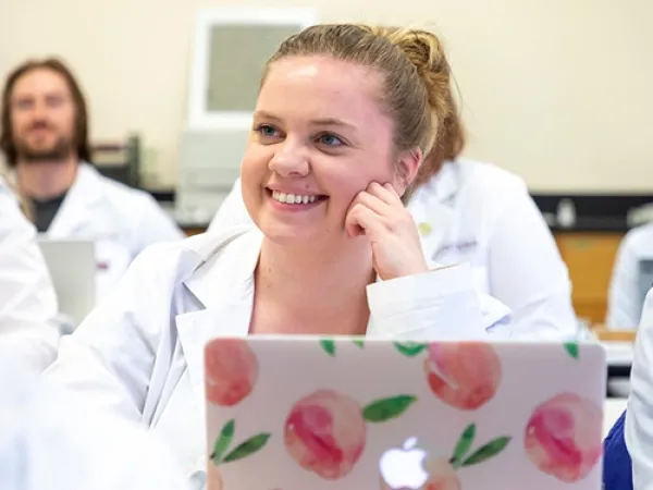 A student wearing a white lab coat listens to an instructor in the classroom