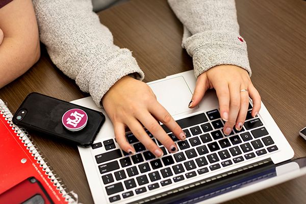 A student works on a laptop. Cell phone with a UM pop socket sits nearby