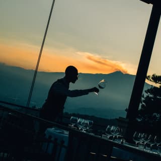 Angled image of a man selecting a wineglass from a table, barely visible in dusk light, with a sunset over Mount Etna behind.