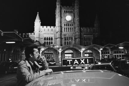 Taxifahrer: A taxi driver smokes a cigarette while waitng for his next fare at Temple Meads Station in Bristol city centre, on 23rd February 1990, in Bristol, England. A 'taxi war' erupted among cab companies vying for control of the taxi business in the Bristol area. Acts of violence between firms have been reported including direct attacks on drivers and vandalism to vehicles.