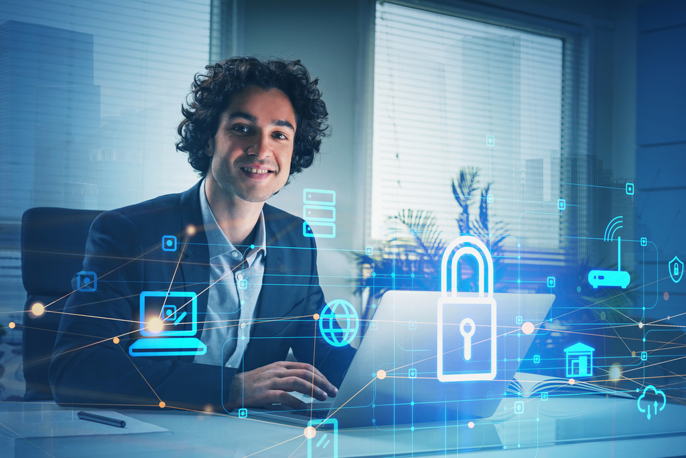 Smiling positive businessman with laptop, desktop at office interior, blue glowing information protection icons. Padlock and business data symbols. Concept of cyber security and data storage