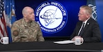 Two men, one in military camouflage and the other in a business suit, are sitting at a desk having a conversation.