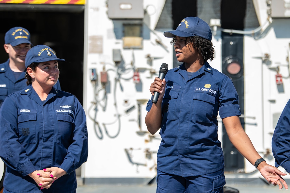 A Coast Guard member is holding a microphone and speaking to other Coast Guard members.