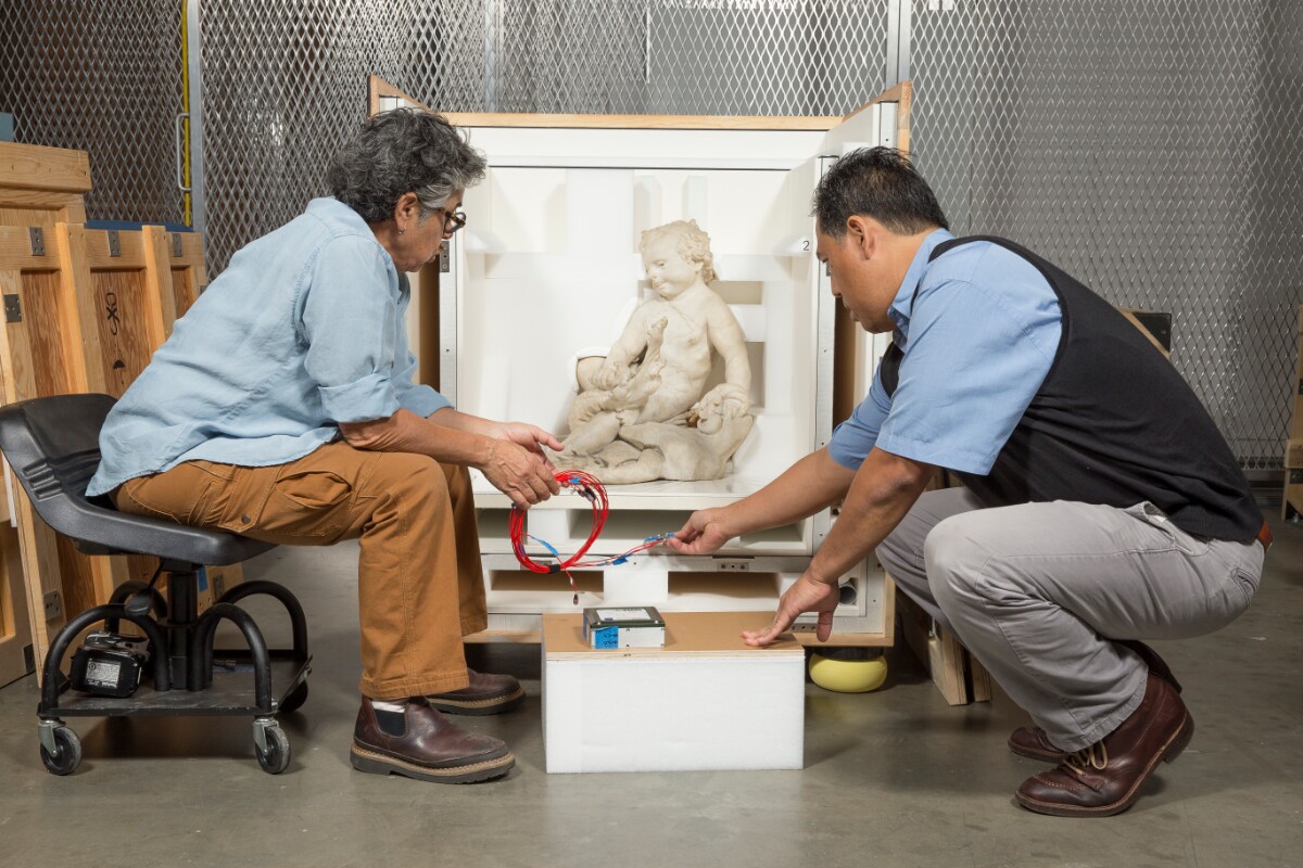 Two people lean in, holding tools towards a wooden crate with a marble sculpture inside 