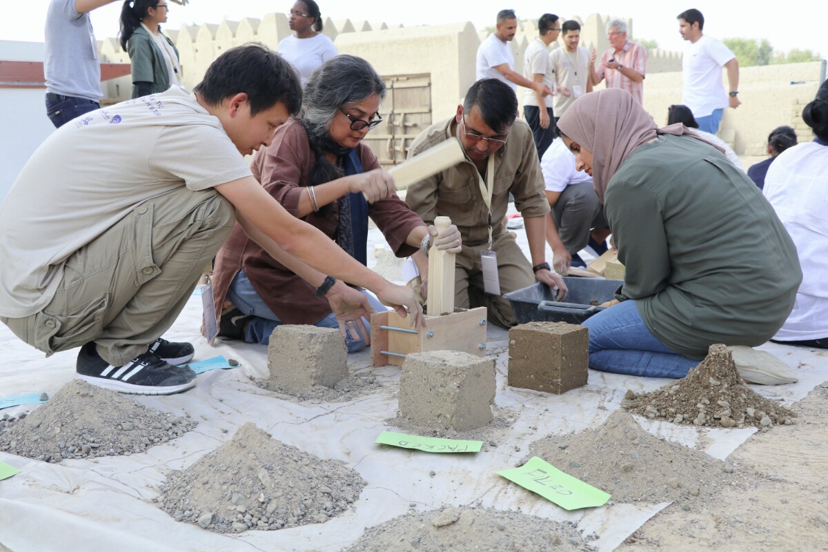 Four people kneel or squat on a tarp on which they make piles of earthen materials into blocks.