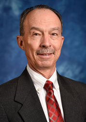 a man with short dark brown hair, wearing a black suit jacket, white shirt and red tie standing in front of a blue background. 