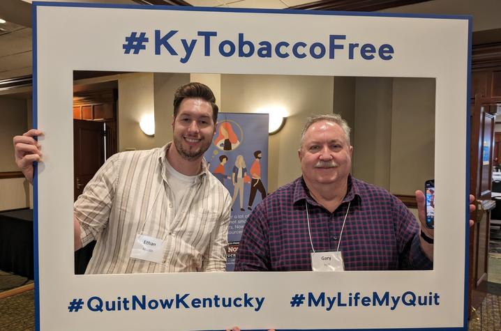 Two men smiling in a frame cutout that says "#KyTobaccoFree #QuitNowKentucky #MyLifeMyQuit"