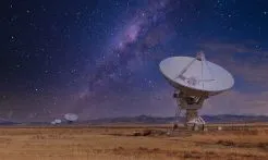 A line of radio dishes on a savannah points to the night sky, with the Milky Way casting a purple glow over mountains.