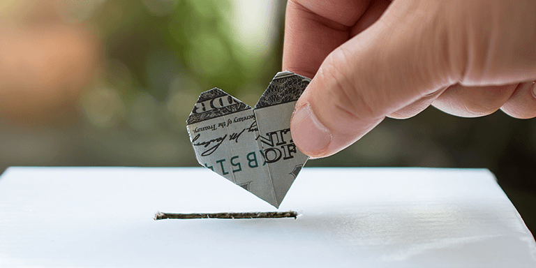 hand holding a dollar bill folded into a heart over a box with a slot to drop money into