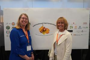 Stacy Clark and Callie Schweitzer stand in front of a banner displaying the International Oak Symposium logo along with smaller logos from the sponsors of the event.