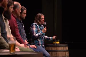 A panel of foresters sit on stage. Tara Keyser has the microphone and is holding a piece of biochar.